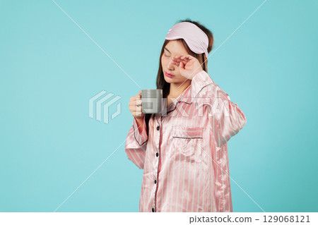 Asian woman in pajama holds a cup after a light sleep, wearing a sleep mask, and relaxing at home, enjoying hot milk for a good mood and restful sleep 129068121