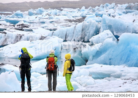 Tourists watching glaciers in Jokulsarlon. Three tourists in brightly colored clothing observe the azure colored floating icebergs on the famous Jokulsarlon glacier lagoon in Iceland 129068453
