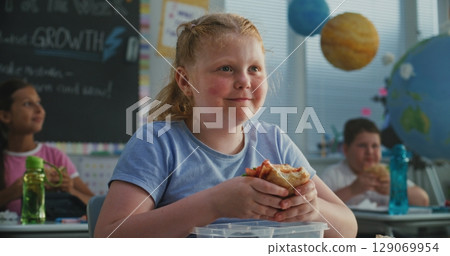 Young Girl Sitting at the Desk, Eating Sandwich from Lunch Box, Talking to Classmate 129069954