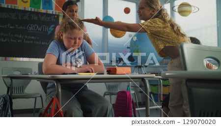 Stressed Primary School Girl Sitting at Desk While Classmates Abusing Her, Throwing Papers 129070020