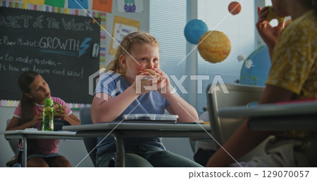 Primary School Girl Sitting at the Desk, Eating Homemade Sandwich, Talking to Classmate 129070057