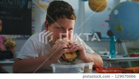 Elementary School Student Sitting at the Desk, Eating Yummy Homemade Sandwich 129070066