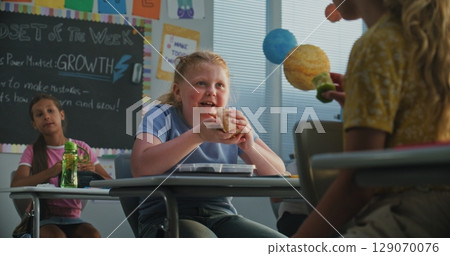 Primary School Girl Sitting at the Desk, Eating Homemade Sandwich, Talking to Classmate Primary School Girl Sitting at the Desk, Eating Homemade Sandwich, Talking to Classmate 129070076