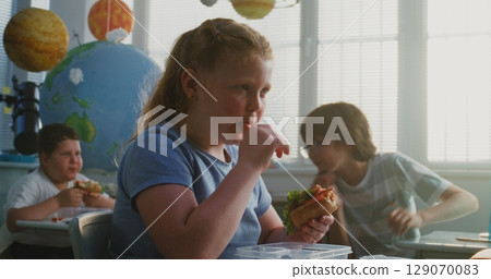 Primary School Girl Eating Delicious Homemade Sandwich from Lunch Box, Talking to Classmates Primary School Girl Eating Delicious Homemade Sandwich from Lunch Box, Talking to Classmates 129070083
