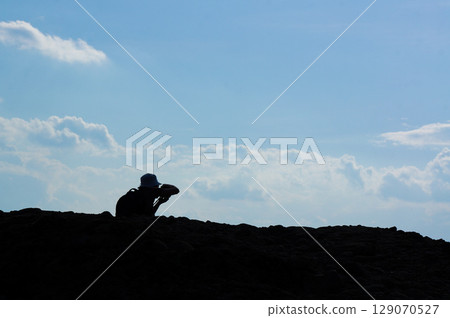 Silhouette of a man sitting on a high rock and taking pictures of the distant scenery 129070527