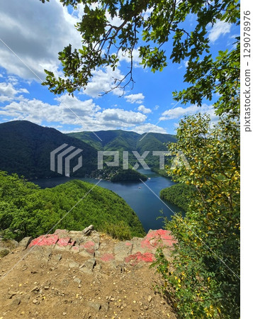 View from Piatra lui Lucaci over the Tarnita lake on a beautiful sunny summer day with blue sky and white clouds; red stone in foreground. Cluj, Romania 129078976