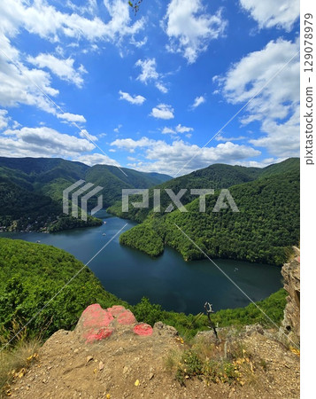 View from Piatra lui Lucaci over the Tarnita lake on a beautiful sunny summer day with blue sky and white clouds; red stone in foreground. Cluj, Romania 129078979