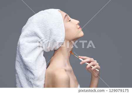 Young asian female with towel wrapped on head applying makeup brush on neck 129079175