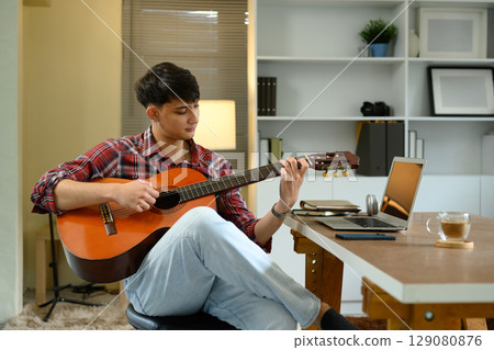 Man playing acoustic guitar while sitting in a modern home office with laptop on the desk 129080876