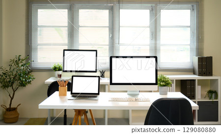 Modern home office with desktop computers and laptop on white desk with blank screens Modern home office with desktop computers and laptop on white desk with blank screens 129080880
