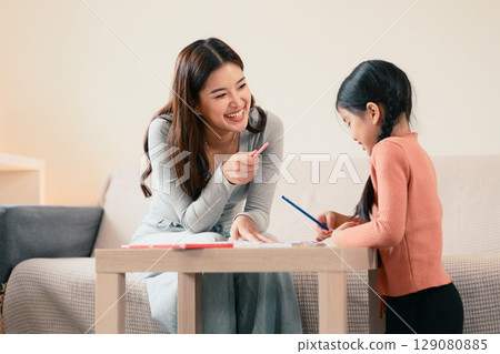 Smiling mother helping her daughter with homework while sitting together at table at home. Smiling mother helping her daughter with homework while sitting together at table at home. 129080885