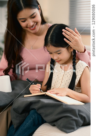 Happy mom supporting her child during homework or creative writing activity on a sofa. Happy mom supporting her child during homework or creative writing activity on a sofa. 129080886