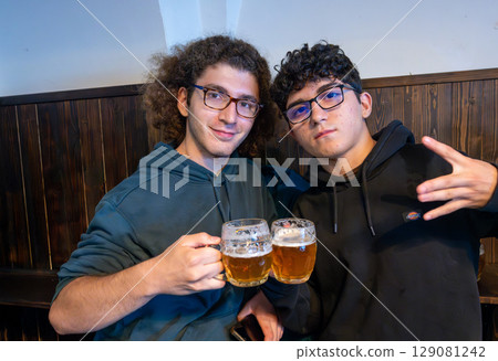 Prague, Czech Republic, August 5, 2023. Two friends look into the camera while toasting in a pub with two mugs of beer. A lifestyle of shared drinking. 129081242