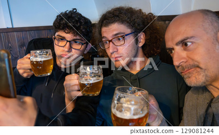 Prague, Czech Republic, August 5, 2023. Group selfie with two young people and a middle-aged man drinking a mug of beer. Lifestyle, drinking together across generations, and the use of technology. Prague, Czech Republic, August 5, 2023. Group selfie with two young people and a middle-aged man drinking a mug of beer. Lifestyle, drinking together across generations, and the use of technology. 129081244