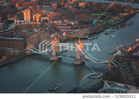 Tower Bridge spans the River Thames in an aerial view, with London's cityscape in the background. The scene is lit by a warm sunset, featuring City Hall and boats. 129083011