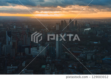 Aerial view of London during sunset with modern skyscrapers, including The Shard, under a sky of orange and yellow hues casting a warm city glow. 129083012