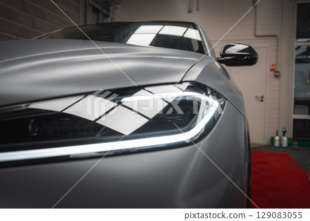 Close up view of the illuminated front left headlight of a Lamborghini Urus with a matte silver finish, set in an indoor garage or showroom. 129083055