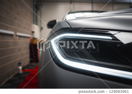 Close up view of a Lamborghini Urus headlight with angular LED design, matte gray finish, and a garage background featuring brick walls and red flooring. 129083061
