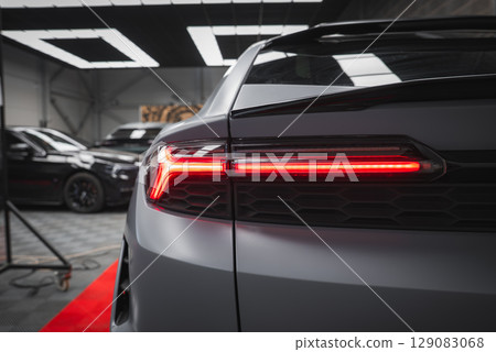 Close up view of a Lamborghini Urus rear taillight illuminated in red, with matte gray finish, honeycomb details, and luxury cars in a showroom setting. 129083068