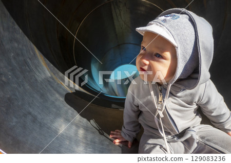Toddler girl with excited and amused expression enjoys crawling in grey slide tube on playground. Child laughs and smiles playing in city park 129083236