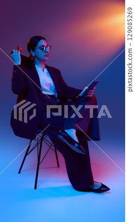 Businesswoman holding smartphone while sitting and playing with her hair under neon light Businesswoman holding smartphone while sitting and playing with her hair under neon light 129085269