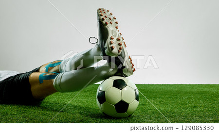 Soccer player resting legs over ball with taped thighs on grass 129085330