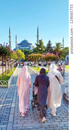 Rear view of women walking towards the Blue Mosque in Istanbul. Three women in muslim clothing walk along a sidewalk surrounded by flowering trees Rear view of women walking towards the Blue Mosque in Istanbul. Three women in muslim clothing walk along a sidewalk surrounded by flowering trees 129085755