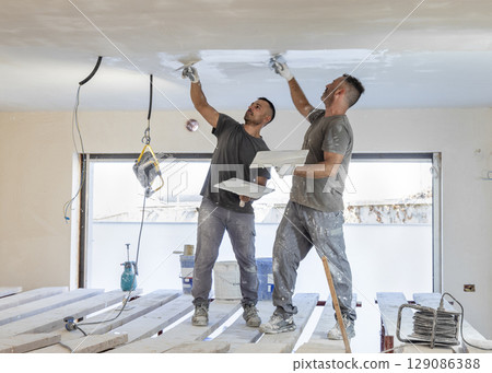 Two men working standing on scaffoldings, use trowel and hawk to plaster ceiling, construction 129086388