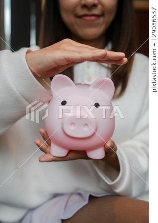 Close-up of a woman holding a pink piggy bank, symbolizing savings and financial security in a cozy home environment. Close-up of a woman holding a pink piggy bank, symbolizing savings and financial security in a cozy home environment. 129086537