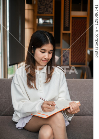 A young woman sits on a sofa, writing in a notebook, surrounded by a modern and cozy interior setting. A young woman sits on a sofa, writing in a notebook, surrounded by a modern and cozy interior setting. 129086541