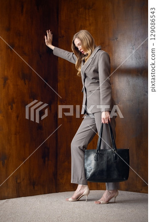 A stylish young woman, long blonde hair, poses in a grey suit, holding a black leather bag against a warm wooden background 129086543