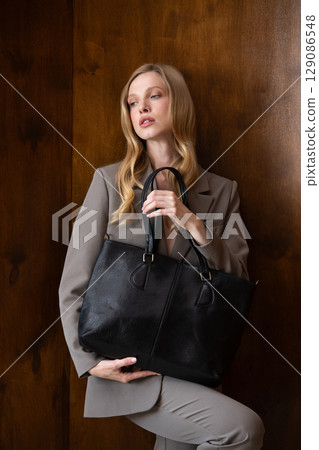 A stylish young woman, long blonde hair, poses in a grey suit, holding a black leather bag against a warm wooden background 129086548