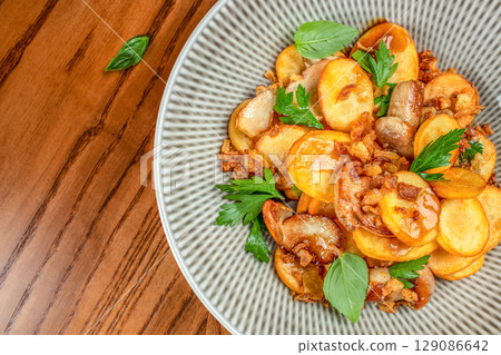 Crispy golden potato slices garnished with herbs and caramelized onions are served in a patterned ceramic bowl on a wooden table. top view. copy space Crispy golden potato slices garnished with herbs and caramelized onions are served in a patterned ceramic bowl on a wooden table. top view. copy space 129086642