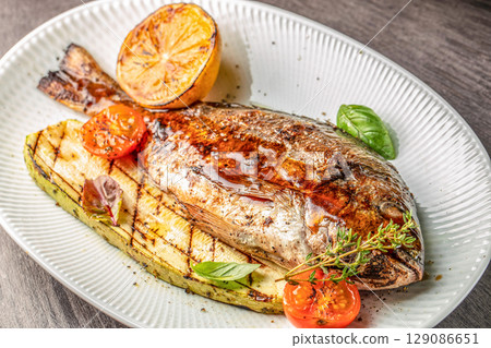 Whole grilled fish served with charred zucchini, cherry tomatoes, and lemon on a white plate. top view 129086651