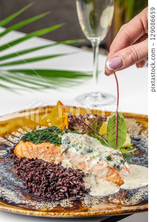 Baked salmon topped with creamy mustard sauce served with black rice and fresh greens on a decorative plate, with a glass of white wine in the background Baked salmon topped with creamy mustard sauce served with black rice and fresh greens on a decorative plate, with a glass of white wine in the background 129086659