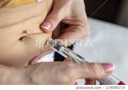Close up shot of the woman with beautiful hands, preparing hormone medicine and injecting herself to the abdomen with pierced bellybutton. 129086714