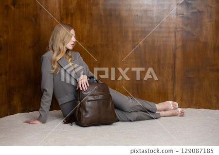 A stylish young woman, long blonde hair, sitting and poses in a grey suit, holding a brown leather backpack against a warm wooden background A stylish young woman, long blonde hair, sitting and poses in a grey suit, holding a brown leather backpack against a warm wooden background 129087185