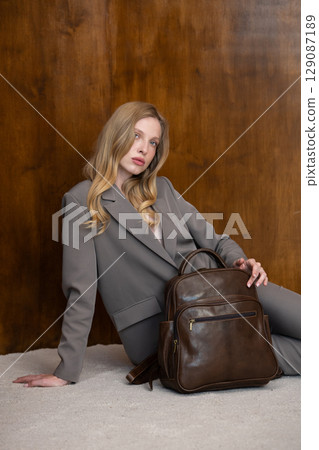 A stylish young woman, long blonde hair, sitting and poses in a grey suit, holding a brown leather backpack against a warm wooden background 129087189