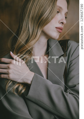 A beautiful young woman with long blonde wavy hair, wearing a stylish grey blazer, gazes thoughtfully to the side against a warm, blurred background A beautiful young woman with long blonde wavy hair, wearing a stylish grey blazer, gazes thoughtfully to the side against a warm, blurred background 129087190