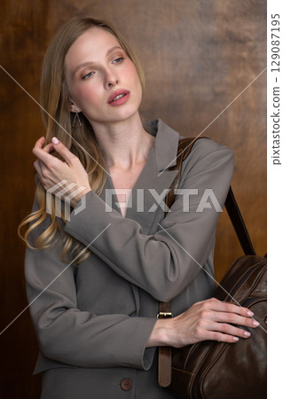 A stylish young woman, long blonde hair, poses in a grey suit, holding a brown leather backpack against a warm wooden background A stylish young woman, long blonde hair, poses in a grey suit, holding a brown leather backpack against a warm wooden background 129087195