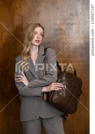 A stylish young woman, long blonde hair, poses in a grey suit, holding a brown leather backpack against a warm wooden background A stylish young woman, long blonde hair, poses in a grey suit, holding a brown leather backpack against a warm wooden background 129087197