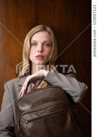 A stylish young woman, long blonde hair, poses in a grey suit, holding a brown leather backpack against a warm wooden background A stylish young woman, long blonde hair, poses in a grey suit, holding a brown leather backpack against a warm wooden background 129087222
