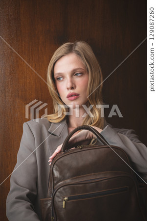 A stylish young woman, long blonde hair, poses in a grey suit, holding a brown leather backpack against a warm wooden background A stylish young woman, long blonde hair, poses in a grey suit, holding a brown leather backpack against a warm wooden background 129087260