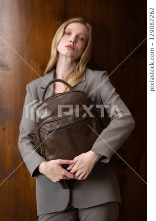 A stylish young woman, long blonde hair, poses in a grey suit, holding a brown leather backpack against a warm wooden background A stylish young woman, long blonde hair, poses in a grey suit, holding a brown leather backpack against a warm wooden background 129087262