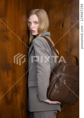 A stylish young woman, long blonde hair, poses in a grey suit, holding a brown leather backpack against a warm wooden background 129087299