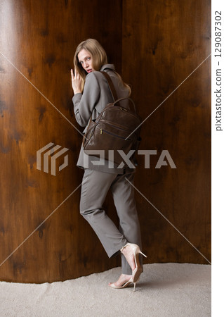 A stylish young woman, long blonde hair, poses in a grey suit, holding a brown leather backpack against a warm wooden background 129087302