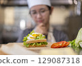 Thoughtful Young Asian Female Chef Evaluating Sandwich in Commercial Kitchen 129087318