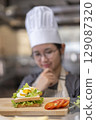 Thoughtful Young Asian Female Chef Evaluating Sandwich in Commercial Kitchen 129087320