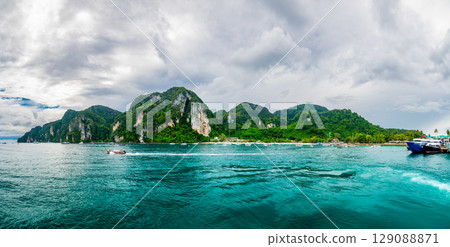 Panoramic wide-angle view of Ko Phi Phi island in Thailand from the sea, surrounded by numerous small and large boats on the clear tropical water under a sunny sky. 129088871