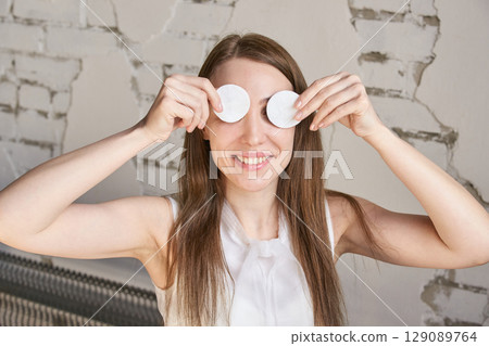 Young caucasian female smiling with cotton pads on eyes against rustic wall 129089764